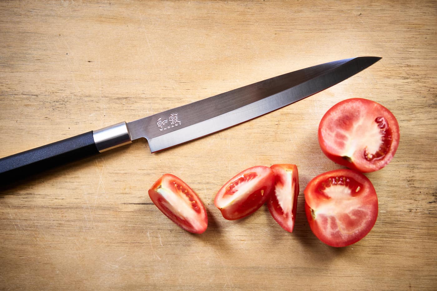 Ein japanisches Yanagiba Messer auf einem Holzbrett, daneben eine geschnittene Tomate.