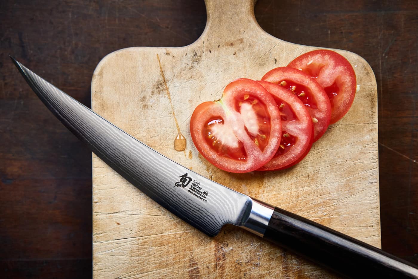 Ein Gokuju Messer mit dunklem Griff auf einem Holzbrett. Daneben eine aufgeschnittene Tomate.