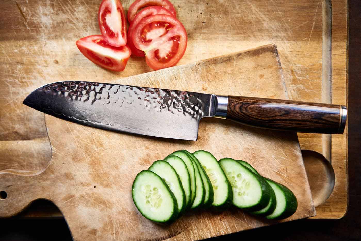 Ein japanisches Santoku Messer auf einem Holzbrett, daneben geschnittene Gurken und Tomaten